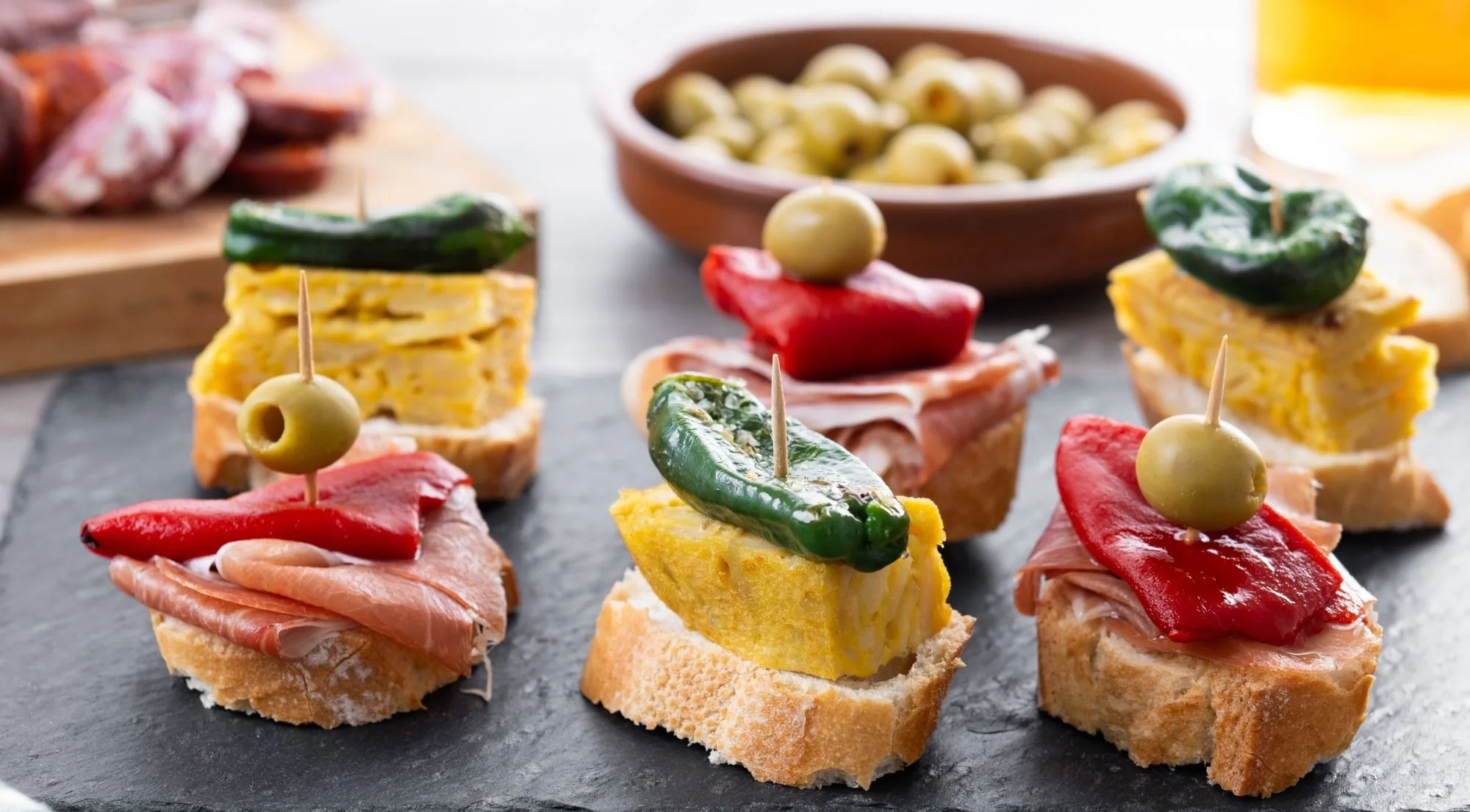 Pintxos lined up on a slate board: tortilla, jamon and piquillo pepper on sliced baguette