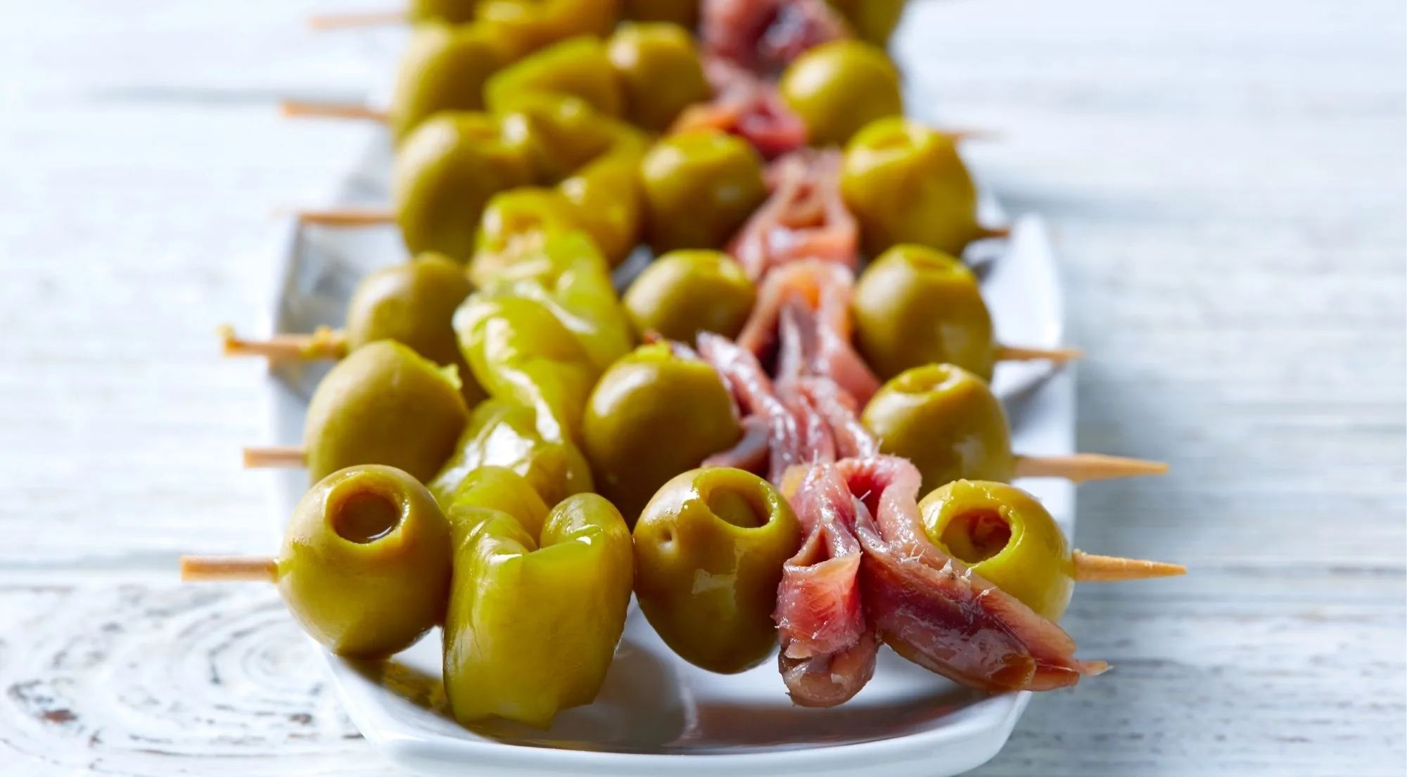 Row of olive, guindilla and anchovy skewers on a white serving plate