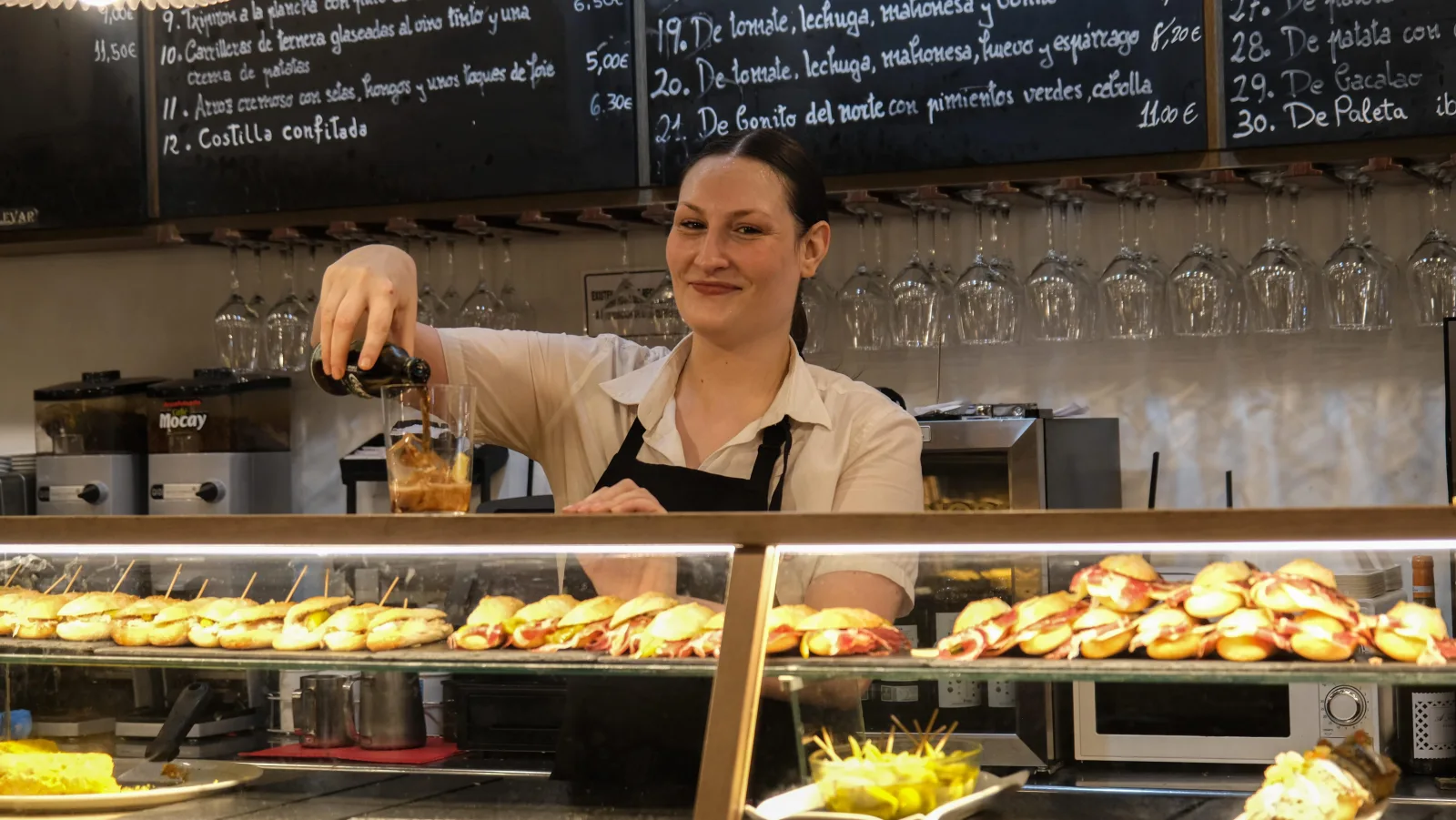 Barmaid pouring a drink behind a counter covered with rows of pintxos
