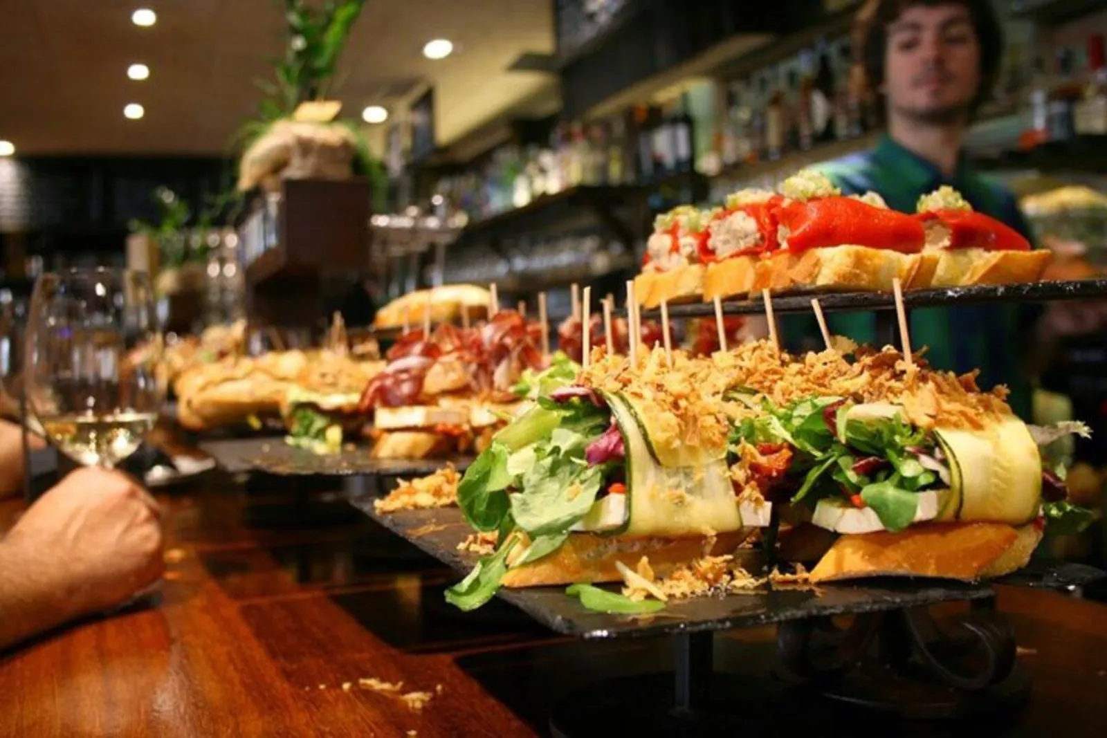 Bar counter stacked with long filled pintxos on toasted bread, barman working behind