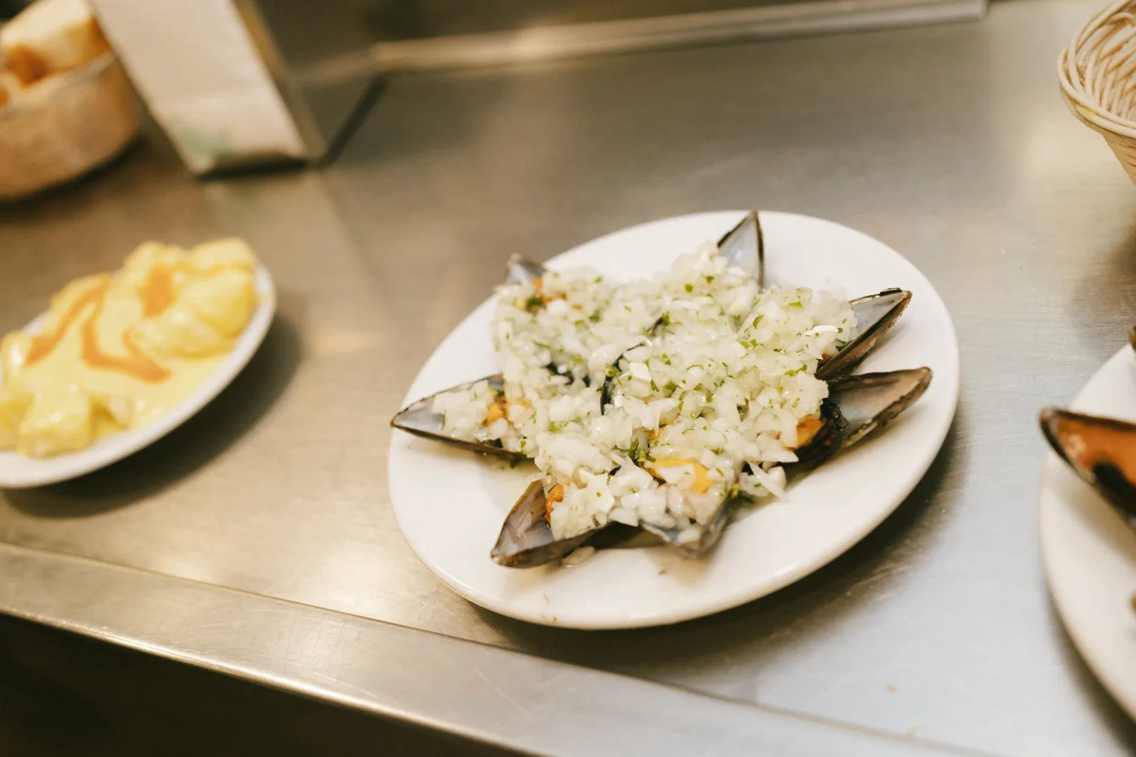 Plate of mussels served with a side of potatoes on a stainless steel bar top