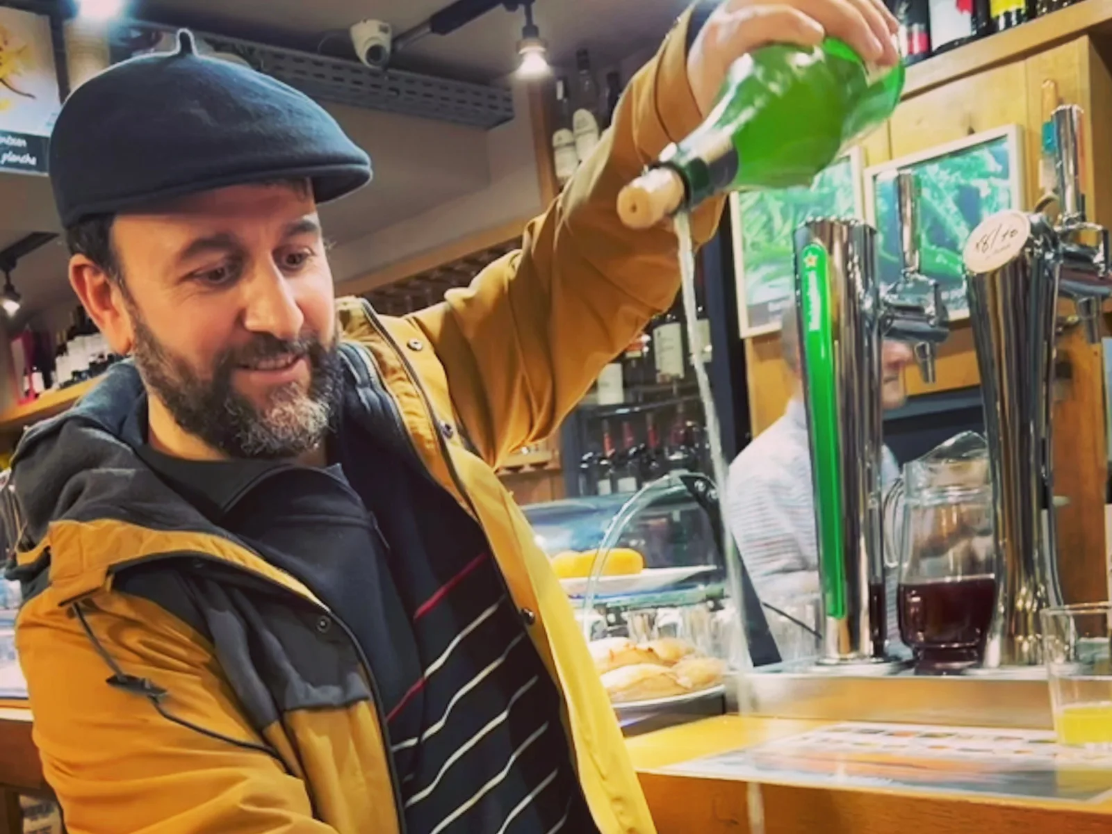 Barman in a Basque beret pouring cider from a green bottle held high above the counter