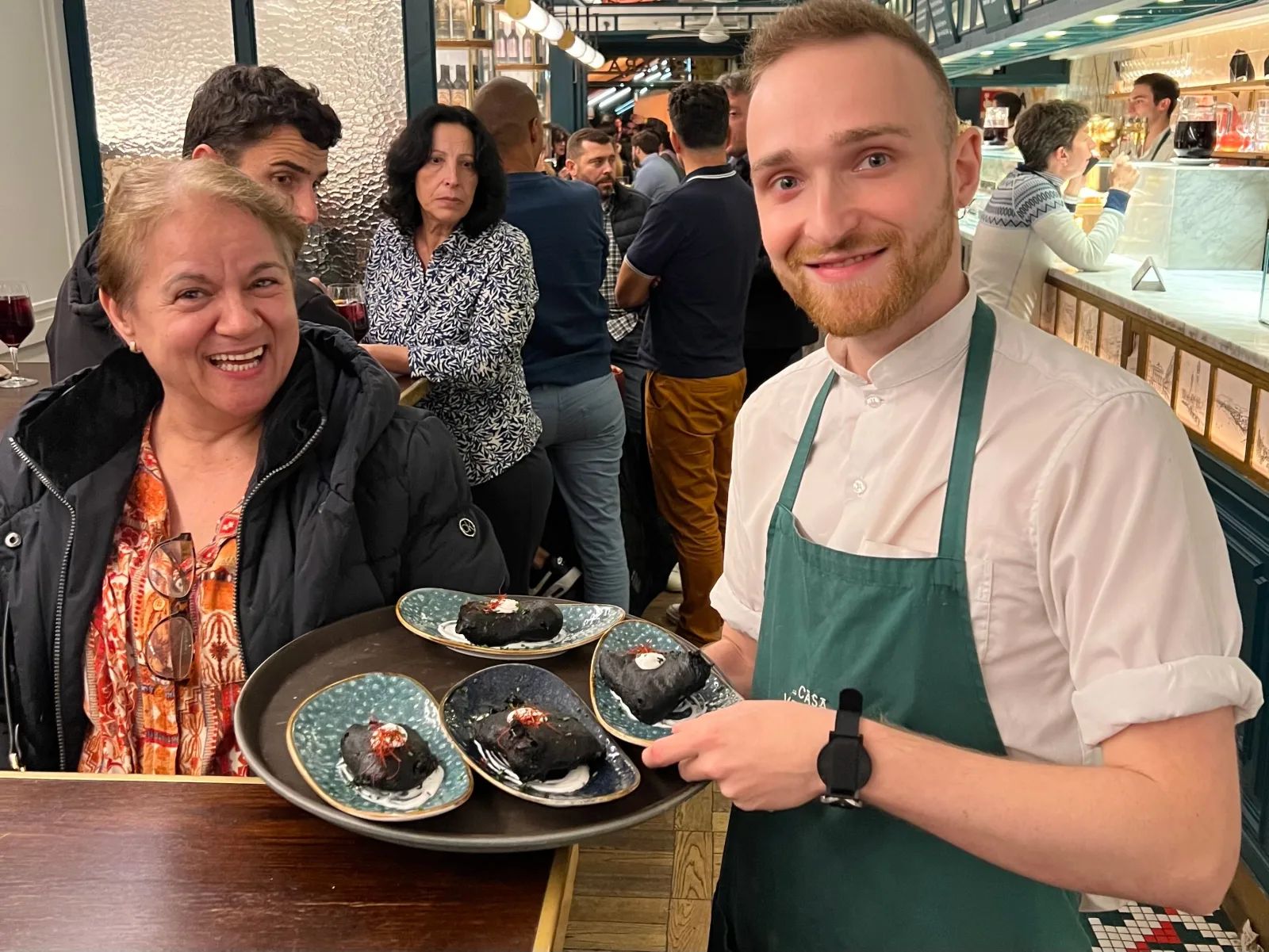 Chef presenting a slate tray of pintxos to a smiling guest inside a market bar