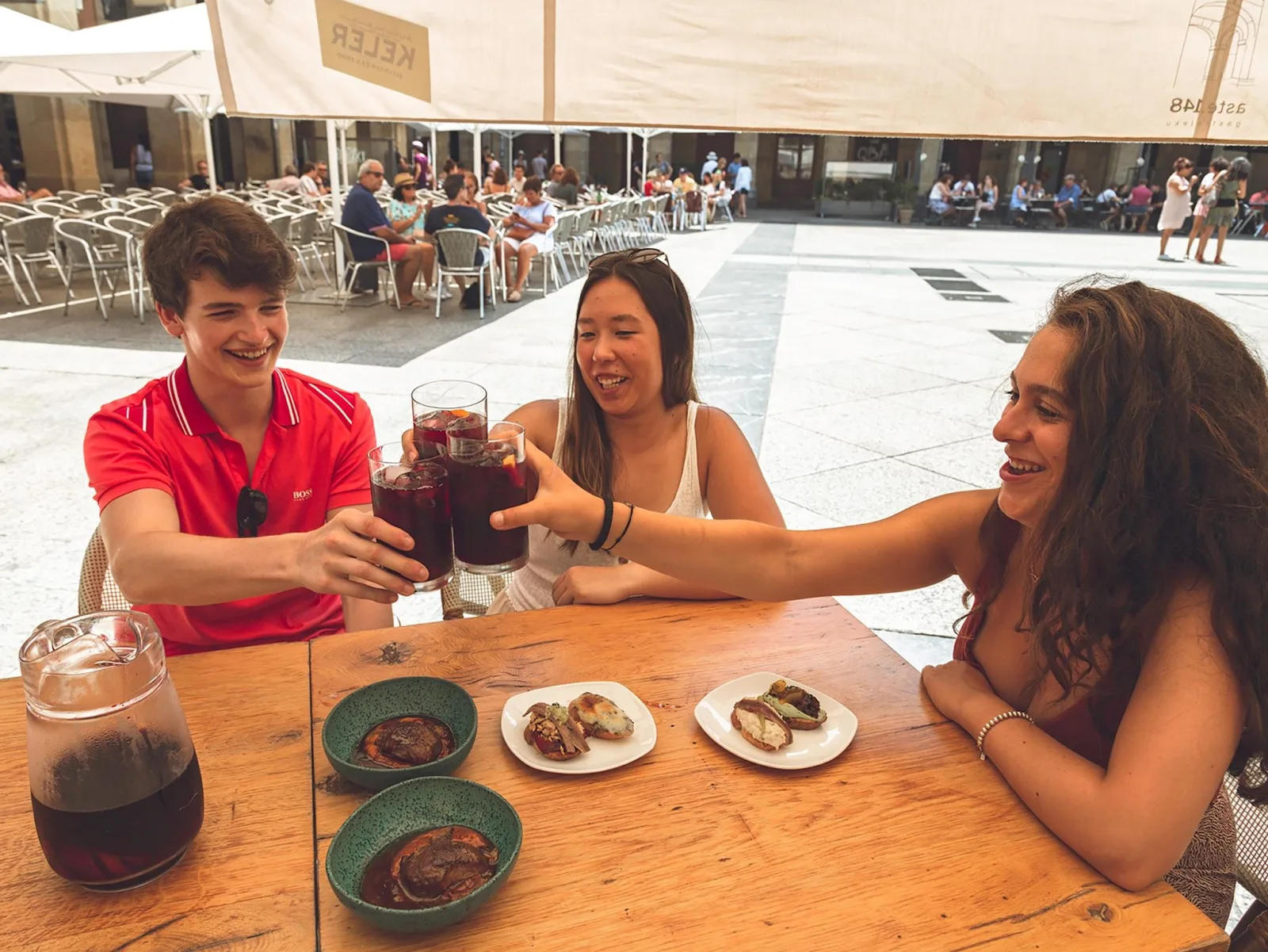 Three guests toasting with red wine over pintxos at an outdoor table on the square