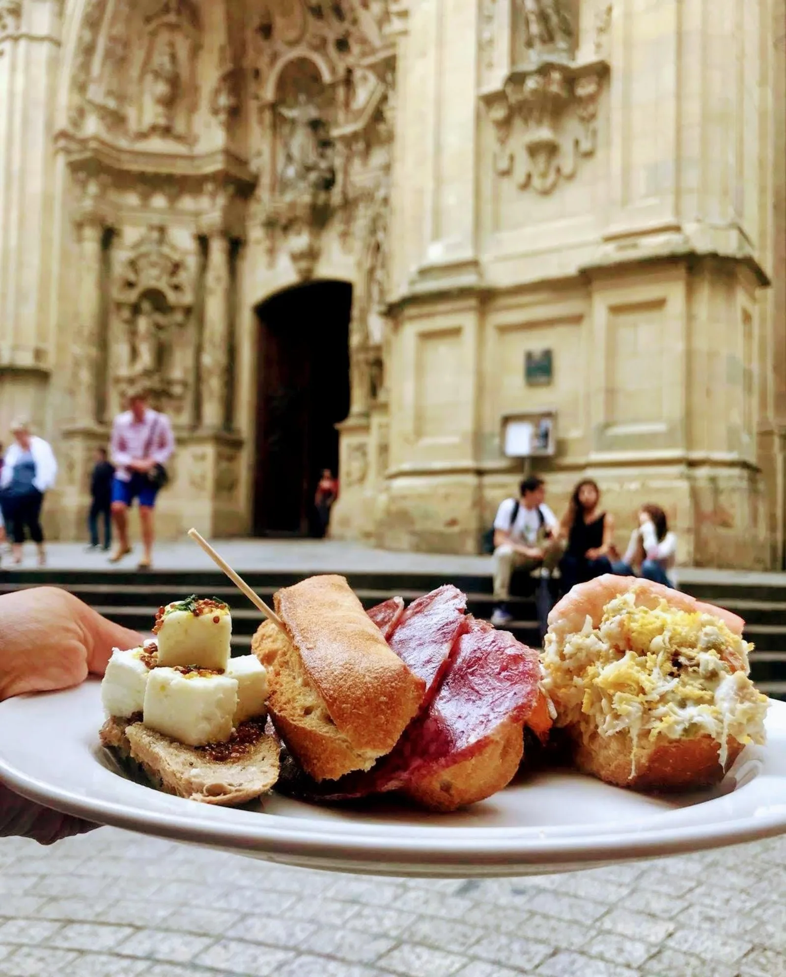 Plate of pintxos held up in front of the carved facade of San Vicente church