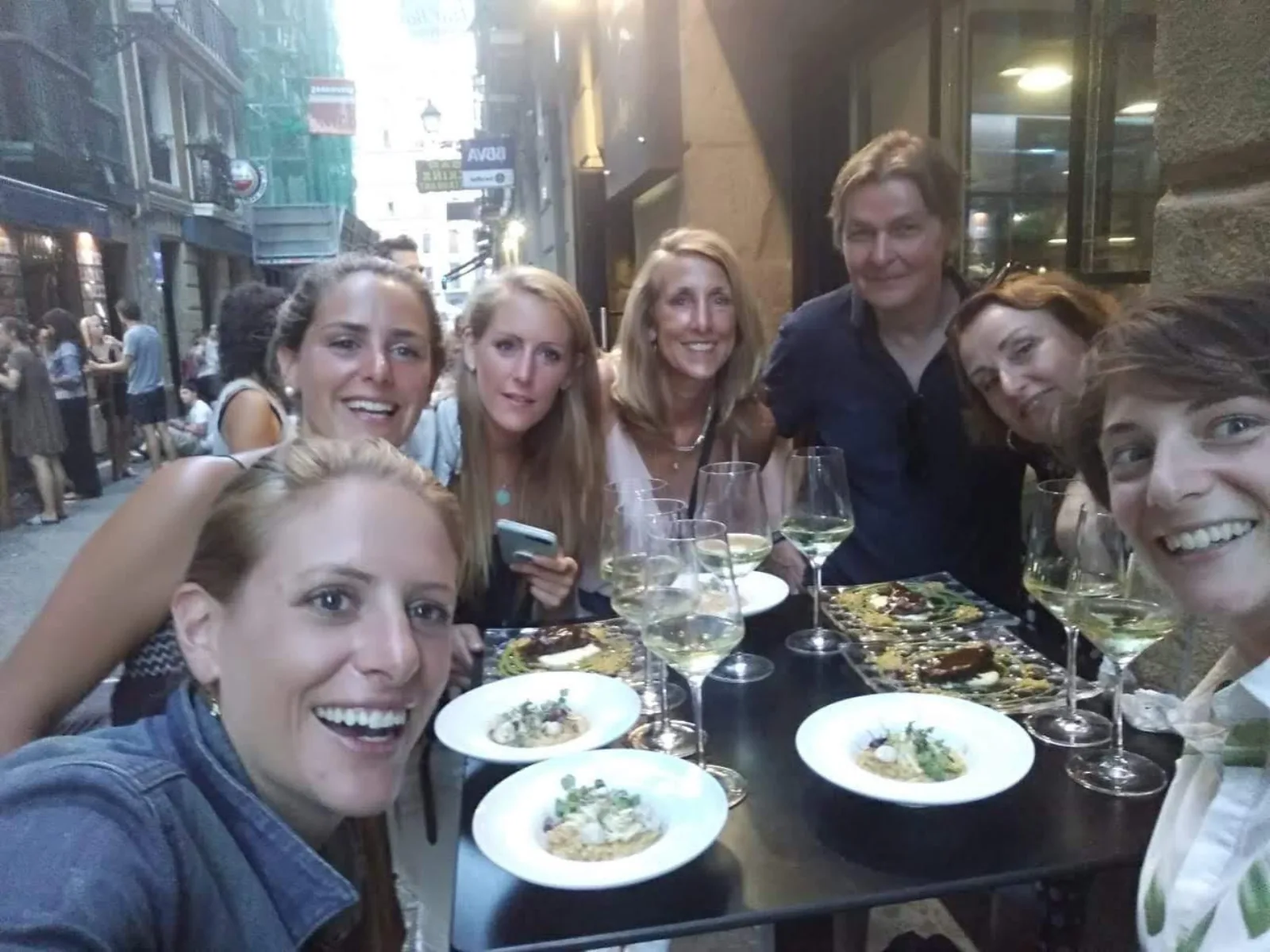 Group photo around a table of white wine and pintxos in a narrow lit Old Town street