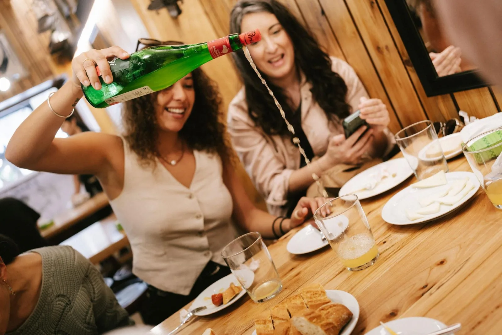 Guest pouring cider from a green bottle held high while the group watches and laughs