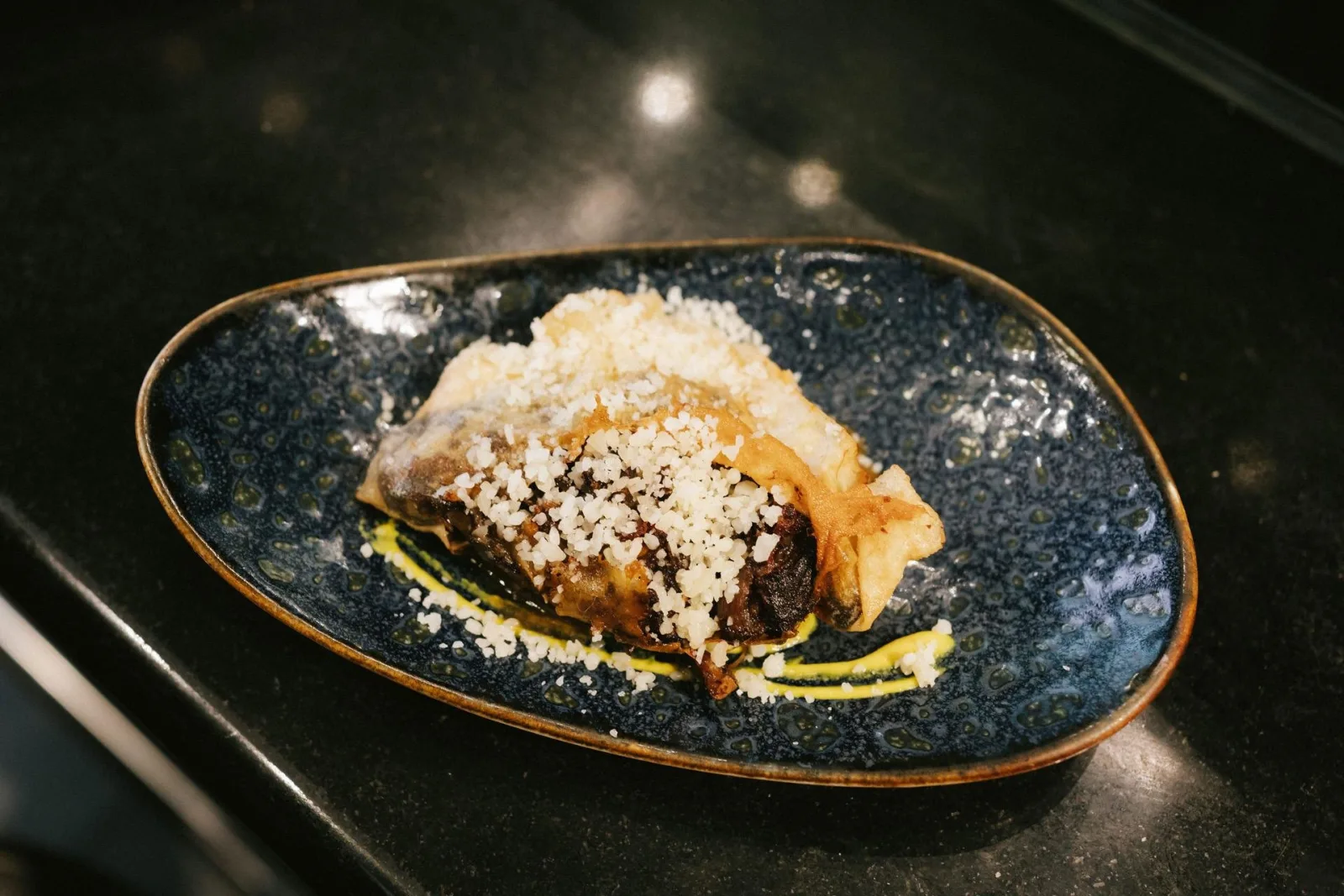 Pintxo of braised meat topped with grated cheese on a dark speckled plate