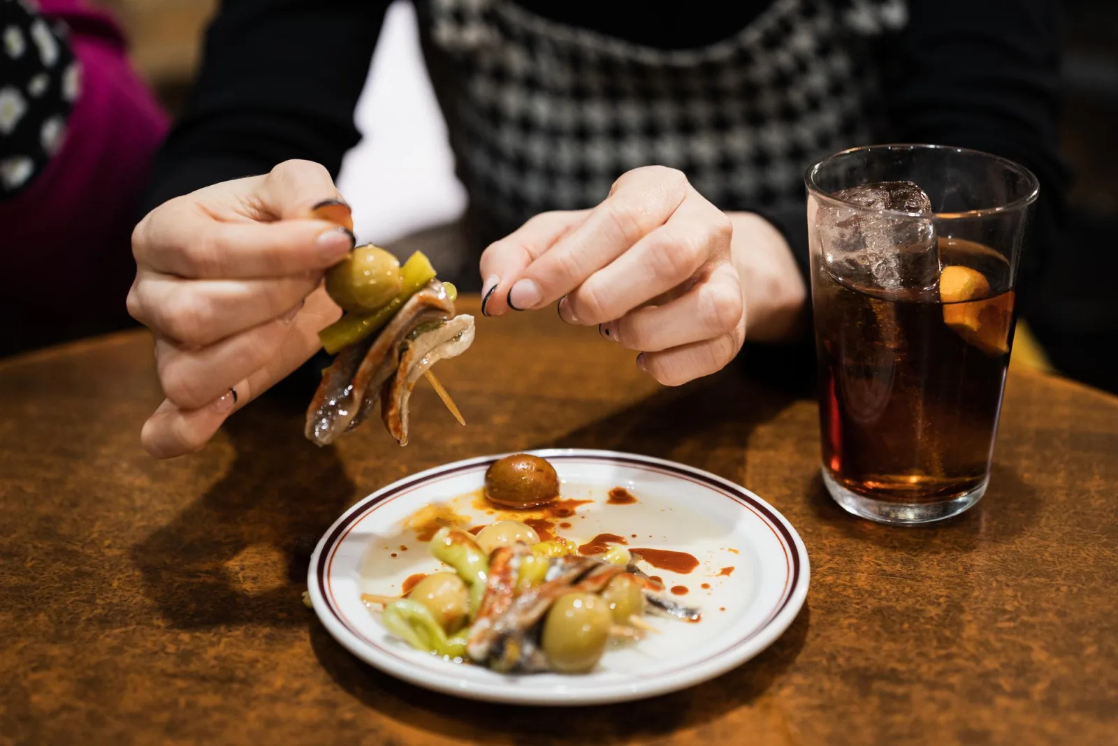 Guest eating a gilda skewer over a plate of pintxos next to a glass of vermouth