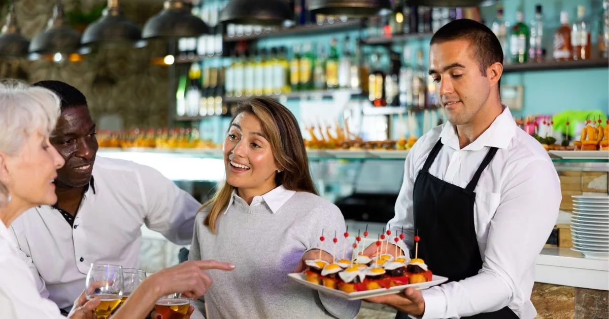 A bartender serving pintxos and drinks to a group at a busy bar counter